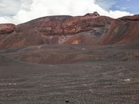 a person with an orange backpack walking on a hill in the mountains with red rocks and grass and dirt and red soil and a white cloud