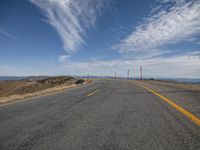 a curve is shown on a deserted road under blue cloudy skies over the mountains and ocean