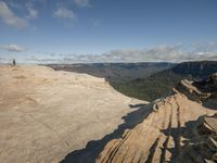 people standing atop a rock and ledge overlooking valley below a blue - tinged sky, below a clear - filled, clear day