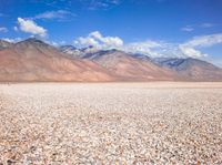 a field in a desert with mountains in the background with clouds and rocks and gravel