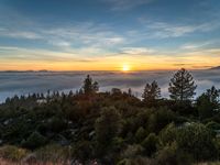 an orange sky above the clouds on a mountain side at sunset with trees and hills below