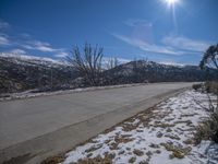 an empty street next to a tree and mountains covered in snow with a sunny sky