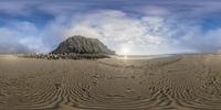 a mountain covered in clouds near the ocean and waves on a beach, with sun peeking above it