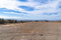 a dirt field that has some trees on it and mountains in the background with clouds