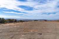 a dirt field that has some trees on it and mountains in the background with clouds