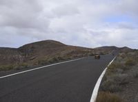 a truck is driving down an asphalt street near mountains, and has two black motorbikes