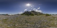 a field with some rocks near a mountain with a clear sky in the background and the sun