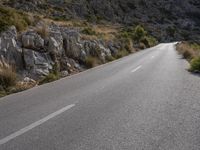 a lone motorcycle is riding in the road beside a rock cliff wall and scrubby