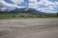 a dirt road running in front of tall mountain range near gate and wood fence with green grass