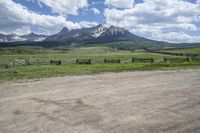 a dirt road running in front of tall mountain range near gate and wood fence with green grass