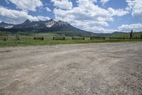 a dirt road running in front of tall mountain range near gate and wood fence with green grass