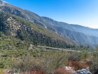 a winding road on the side of a mountain with mountains behind it with a blue sky in the background