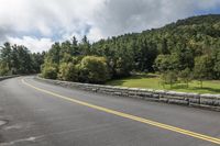 a curve is seen on a bridge over the mountains road in the background are evergreen trees and clouds