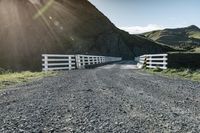 a white gate that is next to a gravel road with mountains in the background with sun flares out