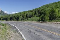 a motorcycle rider rides on the side of a road near a wooded area and mountains