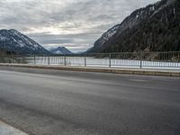 two motorcycles on a long paved highway near a large mountain lake surrounded by snow capped trees