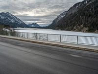 two motorcycles on a long paved highway near a large mountain lake surrounded by snow capped trees