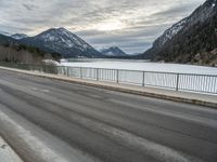 two motorcycles on a long paved highway near a large mountain lake surrounded by snow capped trees