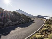 a road running up the side of a mountain near the ocean with sun going through the clouds