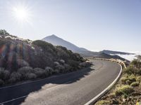 a road running up the side of a mountain near the ocean with sun going through the clouds