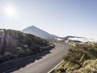 a road running up the side of a mountain near the ocean with sun going through the clouds