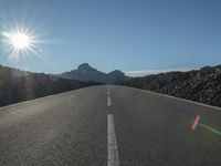 an empty road in the sun with sun flares behind it and a mountain range in the background