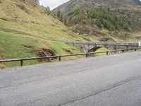 the motorcycle is traveling on a mountain road near a fence and stone bridge over a valley