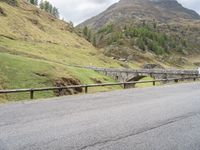 the motorcycle is traveling on a mountain road near a fence and stone bridge over a valley