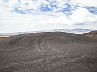 a mountain with dirt mounds on the ground underneath clouds and a blue sky in the distance