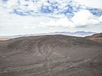 a mountain with dirt mounds on the ground underneath clouds and a blue sky in the distance