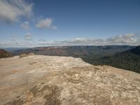 a person sits on the edge of a rock, overlooking mountains and valley beyond on a partly sunny day