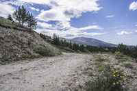 dirt road leading towards green grass and mountain tops and blue sky with clouds above the mountains