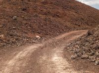 a dirt road winding down to a mountain of rocks, rocks and a cloudy blue sky