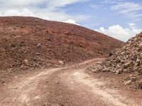 a dirt road winding down to a mountain of rocks, rocks and a cloudy blue sky