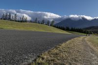 a road with some clouds and some mountains in the background, near some grass and bushes