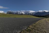 a road with some clouds and some mountains in the background, near some grass and bushes