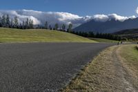 a road with some clouds and some mountains in the background, near some grass and bushes