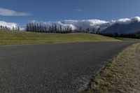 a road with some clouds and some mountains in the background, near some grass and bushes