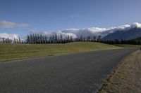 a road with some clouds and some mountains in the background, near some grass and bushes