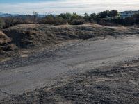 an empty dirt road in front of houses and hills with the sky in the background