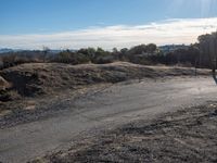 an empty dirt road in front of houses and hills with the sky in the background