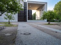 a building sits next to green trees and some gray bricks in the ground on which is a small cobble stone area