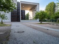 a building sits next to green trees and some gray bricks in the ground on which is a small cobble stone area