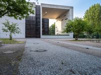 a building sits next to green trees and some gray bricks in the ground on which is a small cobble stone area