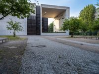 a building sits next to green trees and some gray bricks in the ground on which is a small cobble stone area