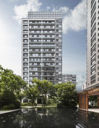 an apartment complex near a lake with trees and plants near the building, there are other buildings in the back ground