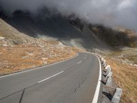 an empty road winds up to the rocky sides of a mountain range as dark clouds hove overhead