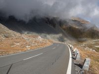 an empty road winds up to the rocky sides of a mountain range as dark clouds hove overhead