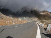 an empty road winds up to the rocky sides of a mountain range as dark clouds hove overhead