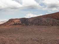 a lone horse standing in the dirt area with a hill behind it, with mountains behind it